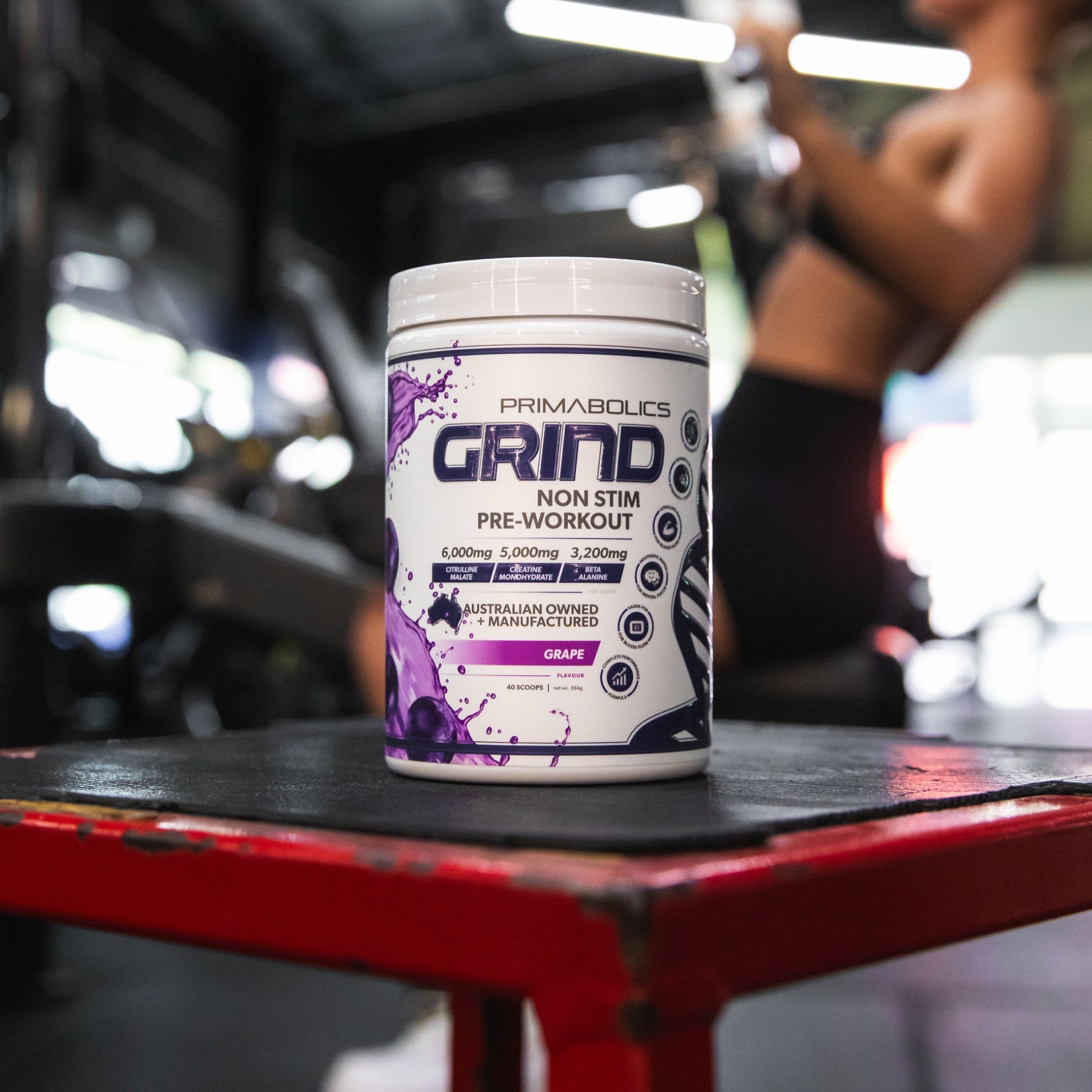 Primabolics Grind pre-workout supplement container on a red table with a blurred background