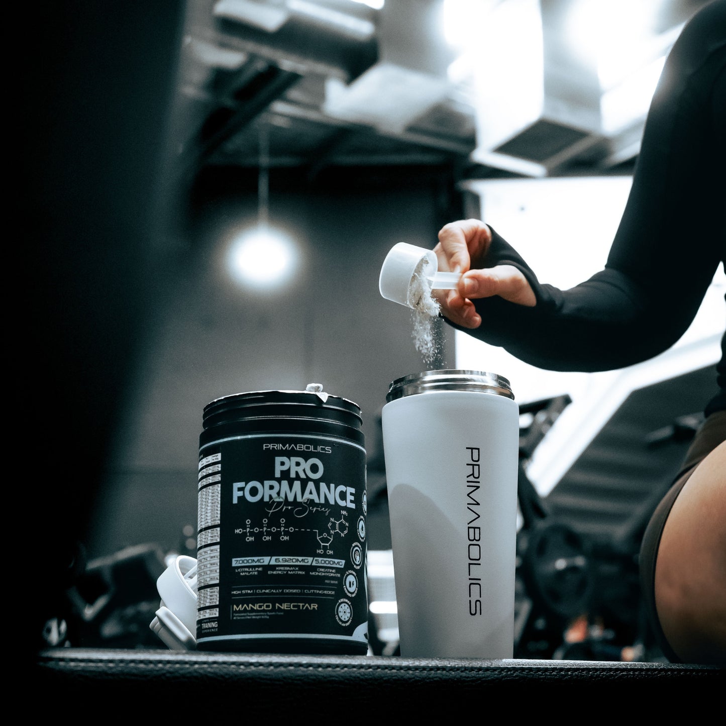 Person pouring a supplement into a Primabolics shaker bottle in a gym setting.